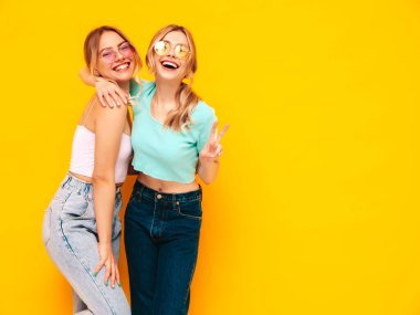 Moda kıyafetleri içinde gülümseyen iki güzel sarışın hippi kadın. Stüdyoda sarı duvarın yanında poz veren kaygısız seksi kadınlar. Pozitif modeller eğleniyor. Neşeli ve mutlu. Güneş gözlükleriyle