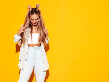 Moda kıyafetleri içinde gülümseyen sarışın güzel bir kadının portresi. Stüdyoda sarı duvarın yanında poz veren kaygısız seksi kadın. Pozitif model içeride eğleniyor. Neşeli ve mutlu