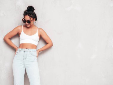 Moda kıyafetleri içinde gülümseyen güzel bir kadın. Stüdyoda gri duvarın yanında poz veren kaygısız seksi kadın. Pozitif esmer model eğleniyor ve deliriyor. Neşeli ve mutlu. Güneş gözlükleriyle
