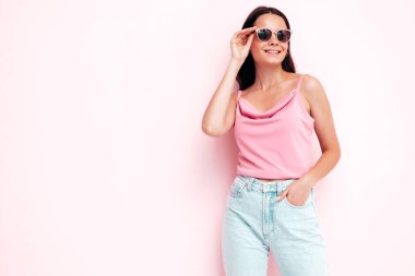 Moda kıyafetleri içinde gülümseyen güzel bir kadın. Stüdyoda pembe duvarın yanında poz veren kaygısız seksi kadın. Pozitif esmer model eğleniyor ve deliriyor. Neşeli ve mutlu. Güneş gözlükleriyle