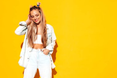 Moda kıyafetleri içinde gülümseyen sarışın güzel bir kadının portresi. Stüdyoda sarı duvarın yanında poz veren kaygısız seksi kadın. Pozitif model içeride eğleniyor. Neşeli ve mutlu