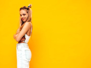 Moda kıyafetleri içinde gülümseyen sarışın güzel bir kadının portresi. Stüdyoda sarı duvarın yanında poz veren kaygısız seksi kadın. Pozitif model içeride eğleniyor. Neşeli ve mutlu