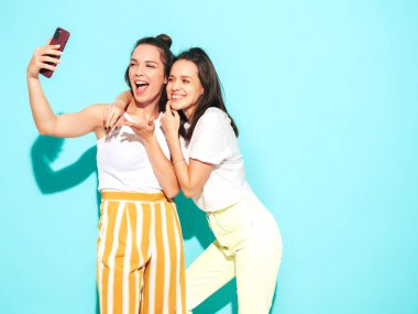 Moda kıyafetleri içinde gülümseyen iki güzel esmer hippi kadının portresi. Mavi duvarın yanında poz veren kaygısız seksi kadınlar. Pozitif modeller eğleniyor. Neşeli ve mutlu. selfie çekmek 