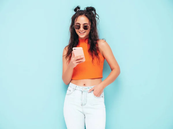 年轻美丽的微笑女性穿着时髦的夏天橙色上衣和牛仔裤衣服 一个性感无忧无虑的女人在工作室的蓝色墙壁附近摆姿势 看着智能手机屏幕 使用手机应用程序的模型 — 图库照片