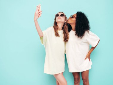 Moda kıyafetleri içinde iki güzel, gülümseyen uluslararası hippi kadın. Stüdyoda mavi duvarın yanında poz veren kaygısız seksi kadınlar. Pozitif modeller güneş gözlükleriyle eğleniyor. Selfie çekiyoruz.