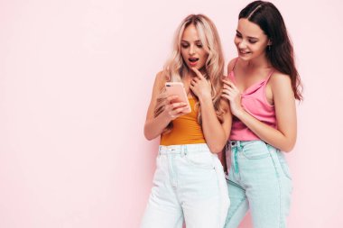 İki güzel, gülümseyen esmer hippi kadın moda yaz kıyafetleri içinde. Stüdyoda pembe duvarın yanında poz veren kaygısız seksi kadınlar. Pozitif modeller eğleniyor. Akıllı telefonu tutmak, cep telefonu uygulamaları kullanmak.