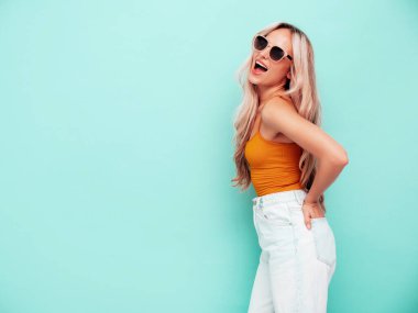 Moda kıyafetleri içinde gülümseyen güzel bir kadının portresi. Stüdyoda mavi duvarın yanında poz veren kaygısız sarışın kadın. Pozitif model içeride eğleniyor. Neşeli ve mutlu. Güneş gözlükleriyle