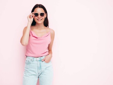 Moda kıyafetleri içinde gülümseyen güzel bir kadın. Stüdyoda pembe duvarın yanında poz veren kaygısız seksi kadın. Pozitif esmer model eğleniyor ve deliriyor. Neşeli ve mutlu. Güneş gözlükleriyle