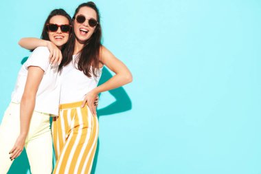 Moda kıyafetleri içinde gülümseyen iki güzel sarışın hippi kadın. Stüdyoda mavi duvarın yanında poz veren kaygısız seksi kadınlar. Pozitif modeller eğleniyor. Neşeli ve mutlu güneş gözlükleriyle