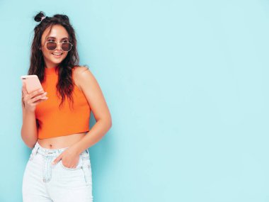 Son moda turuncu tişörtlü ve kot pantolonlu güzel, gülümseyen bir kadın. Stüdyoda mavi duvarın yanında poz veren kaygısız seksi kadın. Akıllı telefon ekranına bakıyorum. Cep telefonu uygulamaları kullanan bir model