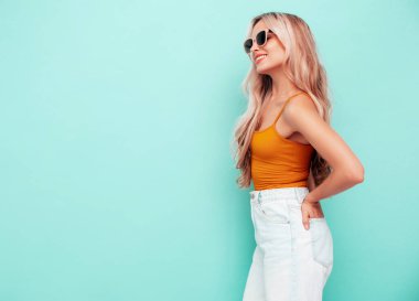 Son moda sarı elbiseli, gülümseyen, güzel sarışın kadın. Stüdyoda mavi duvarın yanında poz veren kaygısız seksi kadın. Pozitif model içeride eğleniyor. Neşeli ve mutlu. Güneş gözlükleriyle