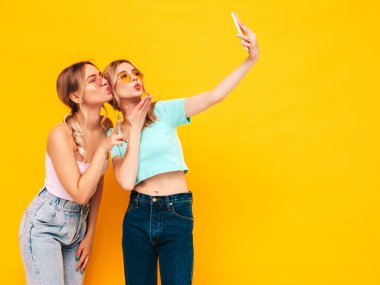 İki güzel, gülümseyen esmer hippi kadın moda yaz kıyafetleri içinde. Stüdyoda sarı duvarın yanında poz veren kaygısız seksi kadınlar. Pozitif modeller eğleniyor. Neşeli ve mutlu. Selfie çekiyorum. 