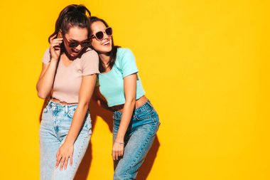 İki güzel, gülümseyen esmer hippi kadın moda yaz kıyafetleri içinde. Stüdyoda sarı duvarın yanında poz veren kaygısız seksi kadınlar. Pozitif modeller eğleniyor. Neşeli ve mutlu