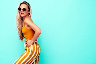 Son moda sarı elbiseli, gülümseyen, güzel sarışın kadın. Stüdyoda mavi duvarın yanında poz veren kaygısız seksi kadın. Pozitif model içeride eğleniyor. Neşeli ve mutlu. Güneş gözlükleriyle