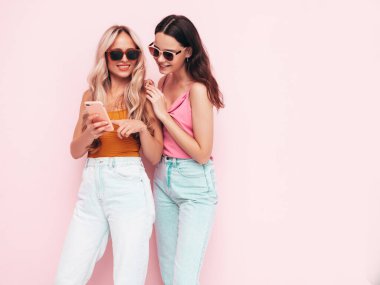 İki güzel, gülümseyen esmer hippi kadın moda yaz kıyafetleri içinde. Stüdyoda pembe duvarın yanında poz veren kaygısız seksi kadınlar. Pozitif modeller eğleniyor. Akıllı telefonu tutmak, cep telefonu uygulamaları kullanmak.