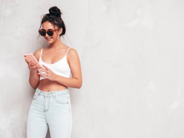 Moda kıyafetleri içinde gülümseyen güzel bir kadın. Stüdyoda gri duvarın yanında poz veren kaygısız seksi kadın. Pozitif esmer model eğleniyor ve deliriyor. Akıllı telefon uygulamaları kullanarak. Telefon bekletiliyor