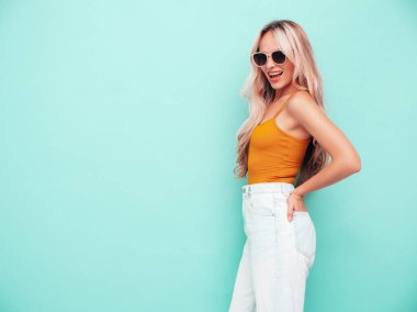 Son moda sarı elbiseli, gülümseyen, güzel sarışın kadın. Stüdyoda mavi duvarın yanında poz veren kaygısız seksi kadın. Pozitif model içeride eğleniyor. Neşeli ve mutlu. Güneş gözlükleriyle