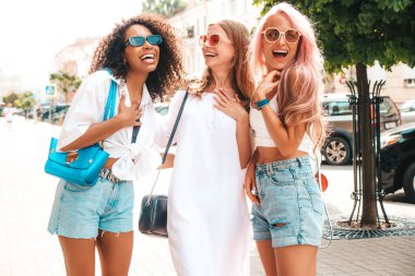 Son moda yaz kıyafetleri içinde gülümseyen üç genç bayan. Sokakta poz veren kaygısız, çok ırklı kadınlar. Güneş gözlükleriyle eğlenen pozitif modeller. Neşeli ve mutlu