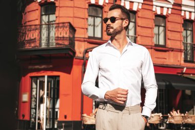 Yakışıklı, kendine güvenen, stil sahibi, lamberseksüel modelin portresi. Beyaz gömlek ve pantolon giymiş modern bir adam. Moda erkeği gün batımında Avrupa 'da sokak arkasındaymış gibi poz veriyor. Güneş gözlükleriyle