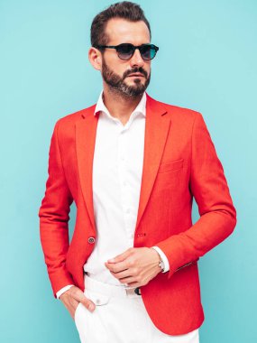 Yakışıklı, kendine güvenen, stil sahibi, lamberseksüel modelin portresi. Zarif kırmızı takım elbise giymiş modern bir adam. Moda erkeği stüdyoda mavi duvarın yanında güneş gözlüklü poz veriyor.