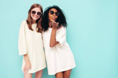 Moda kıyafetleri içinde iki güzel, gülümseyen uluslararası hippi kadın. Stüdyoda mavi duvarın yanında poz veren kaygısız seksi kadınlar. Pozitif modeller eğleniyor. Arkadaşlık kavramı. Güneş gözlükleriyle