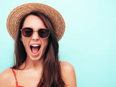 Son moda kırmızı elbisesi içinde gülümseyen güzel bir kadın. Stüdyoda mavi duvarın yanında poz veren kaygısız seksi kadın. Pozitif esmer model şapka ve güneş gözlükleriyle eğleniyor. Çığlıklar ve bağırışlar