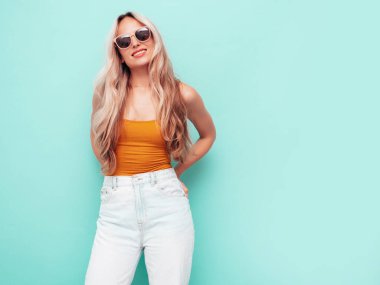 Son moda sarı elbiseli, gülümseyen, güzel sarışın kadın. Stüdyoda mavi duvarın yanında poz veren kaygısız seksi kadın. Pozitif model içeride eğleniyor. Neşeli ve mutlu. Güneş gözlükleriyle