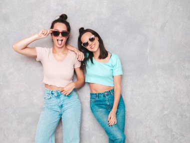 Moda kıyafetleri içinde gülümseyen iki güzel sarışın hippi kadın. Stüdyoda gri duvarın yanında poz veren kaygısız seksi kadınlar. Pozitif modeller eğleniyor. Neşeli ve mutlu. Güneş gözlükleriyle