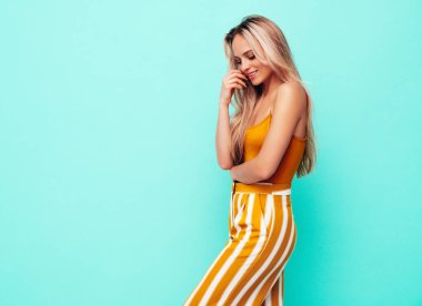 Son moda sarı elbiseli, gülümseyen, güzel sarışın kadın. Stüdyoda mavi duvarın yanında poz veren kaygısız seksi kadın. Pozitif model içeride eğleniyor. Neşeli ve mutlu