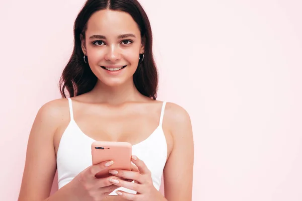 穿着时髦夏装的年轻漂亮微笑的女性 一个性感无忧无虑的女人在工作室的粉红墙边摆姿势 正面的黑发模特看着手机屏幕 持有智能手机和使用应用程序 — 图库照片