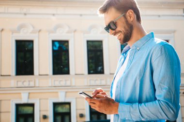 Yakışıklı, güler yüzlü, stil sahibi, lamberseksüel model. Mavi gömlekli, modern bir adam. Moda erkeği sokakta poz veriyor. Gün batımında dışarıda. Akıllı telefon uygulamaları kullanarak. Güneş gözlükleriyle