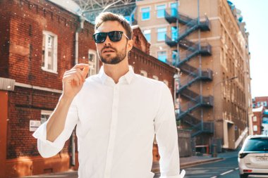 Yakışıklı, kendine güvenen, stil sahibi, Lambersex mankeni portresi. Beyaz gömlek giymiş modern bir adam. Sokakta güneş gözlükleriyle poz veren modacı erkek. Gün batımında dışarıda. 
