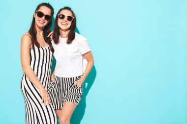 Moda kıyafetleri içinde gülümseyen iki güzel sarışın hippi kadın. Stüdyoda mavi duvarın yanında poz veren kaygısız seksi kadınlar. Pozitif modeller eğleniyor. Neşeli ve mutlu. Güneş gözlükleriyle