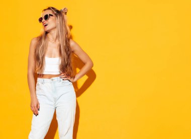 Moda kıyafetleri içinde gülümseyen sarışın güzel bir kadının portresi. Stüdyoda sarı duvarın yanında poz veren kaygısız seksi kadın. Pozitif model içeride eğleniyor. Neşeli ve mutlu güneş gözlükleriyle
