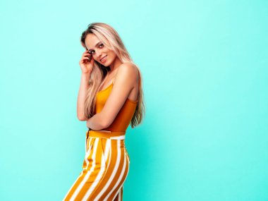 Son moda sarı elbiseli, gülümseyen, güzel sarışın kadın. Stüdyoda mavi duvarın yanında poz veren kaygısız seksi kadın. Pozitif model içeride eğleniyor. Neşeli ve mutlu