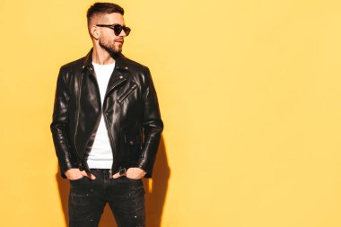 Yakışıklı, kendinden emin Lambersexual modelin portresi. Motosikletli deri ceket ve siyah kot giyen seksi şık bir adam. Moda hippi erkek. Stüdyoda güneş gözlüklü sarı duvarın yanında poz veriyor.