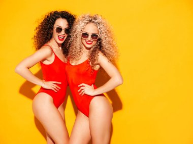 Kırmızı yaz mayo giymiş, gülümseyen iki güzel hippi kadın. Saç stili kıvırcık saçlı moda modeller stüdyoda eğleniyor. Güneş gözlüklü, sarı duvarın yanında poz veren seksi bir kadın.