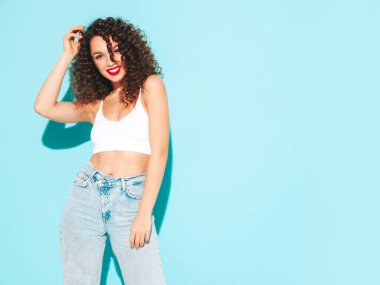 Yeni moda hippi kıyafetleri içinde gülümseyen güzel bir kadın. Stüdyoda mavi duvarın yanında poz veren kaygısız seksi kadın. Afro kıvırcık saçlı esmer bir model. Neşeli ve mutlu