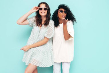 Moda kıyafetleri içinde iki güzel, gülümseyen uluslararası hippi kadın. Stüdyoda mavi duvarın yanında poz veren kaygısız seksi kadınlar. Pozitif modeller eğleniyor. Arkadaşlık kavramı