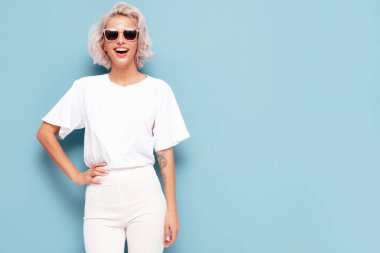 Moda kıyafetleri içinde gülümseyen güzel bir kadın. Stüdyoda mavi duvarın yanında poz veren kaygısız seksi kadın. Pozitif sarışın model eğleniyor ve deliriyor.