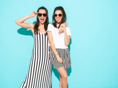 Moda kıyafetleri içinde gülümseyen iki güzel sarışın hippi kadın. Stüdyoda mavi duvarın yanında poz veren kaygısız seksi kadınlar. Pozitif modeller eğleniyor. Neşeli ve mutlu. Güneş gözlükleriyle