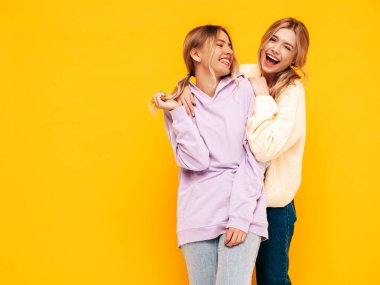 İki güzel, gülümseyen esmer hippi kadın moda yaz kıyafetleri içinde. Stüdyoda sarı duvarın yanında poz veren kaygısız kadınlar. Pozitif modeller eğleniyor. Neşeli ve mutlu