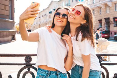 Son moda beyaz tişörtlü ve kot pantolonlu iki güzel gülümseyen yeniyetme kadın. Arka planda poz veren kaygısız kadınlar. Pozitif mankenler eğleniyor, sarılıyor.