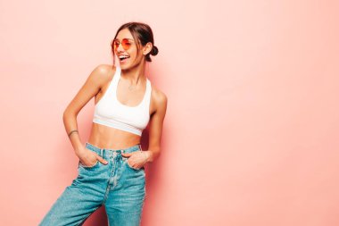 Beyaz formalı bluz ve kot pantolon giymiş güzel bir kadın. Seksi kaygısız neşeli model içeride eğleniyor. Stüdyoda pembe duvarın yanında poz veren sevimli ve pozitif kadın. Güneş gözlükleriyle