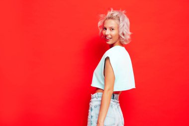 Moda kıyafetleri içinde gülümseyen güzel bir kadın. Stüdyoda kırmızı duvarın yanında poz veren kaygısız bir kadın. Pozitif sarışın model eğleniyor ve gidiyor. Neşeli ve mutlu