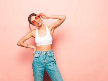 Beyaz formalı bluz ve kot pantolon giymiş güzel bir kadın. Seksi kaygısız neşeli model içeride eğleniyor. Stüdyoda pembe duvarın yanında poz veren sevimli ve pozitif kadın. Güneş gözlükleriyle