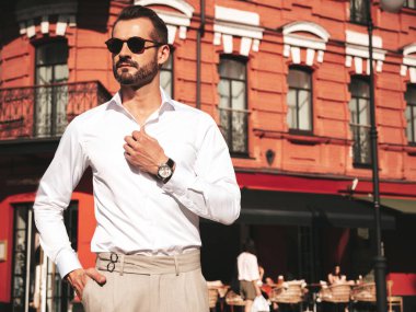 Yakışıklı, kendine güvenen, stil sahibi, lamberseksüel modelin portresi. Beyaz gömlek ve pantolon giymiş modern bir adam. Moda erkeği gün batımında Avrupa 'da sokak arkasındaymış gibi poz veriyor. Güneş gözlükleriyle