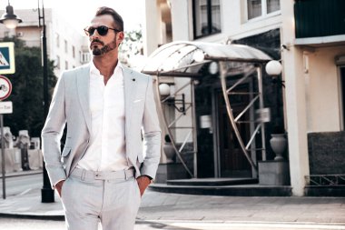 Yakışıklı, kendine güvenen, stil sahibi, lamberseksüel modelin portresi. Zarif beyaz takım elbise giymiş modern bir adam. Moda erkeği gün batımında Avrupa şehrinin arka planında poz veriyor. Güneş gözlükleriyle
