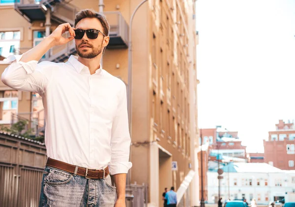 Yakışıklı, gülümseyen stil sahibi Lambersex modelinin portresi. Beyaz gömlek giymiş modern bir adam. Sokakta güneş gözlükleriyle poz veren modacı erkek. Gün batımında dışarıda. 
