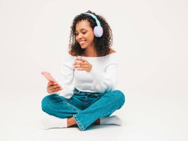 Saçları kıvırcık, saçları kıvırcık, süveter ve kot giyen, kablosuz kulaklıkla müzik dinleyen kaygısız bir kadın. Stüdyoda beyaz arka planda poz veriyor. Akıllı telefon tutuyor.